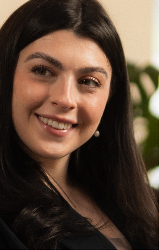A woman with long dark hair smiling, wearing a black top and pearl drop earrings, with a blurred green plant in the background.